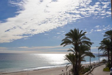 Palmiye ağaçlarıyla dolu bir kumsal, birkaç yüksek bulut ve güneşli bir gün, sakin mavi bir deniz ve gezinti güvertesinin yanında beyaz bir kum.