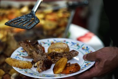 Bir elinde kazıyıcı, diğer elinde neredeyse yemek dolu bir tabakla yemek pişirir ve rostoyu dağıtır.