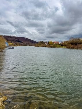 Bulutlu gökyüzü ve uzak tepeleri olan sakin bir nehir, köy kıyı şeridi. Yüksek kalite fotoğraf