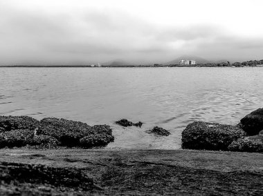 Bulutlu bir gökyüzünün altında kayalar ve arka planda dağlı binalar olan sakin denizin siyah beyaz fotoğrafı.