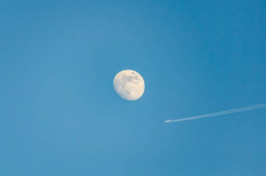 Açık mavi gökyüzünde parlak ayın yanından geçen bir uçak. Telefoto çekimi, dinamik hareket, barışçıl atmosfer, minimalist kompozisyon, açık gökyüzü, havacılık teması, gök kontrastı, seyahat ilhamı.