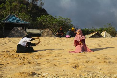 Gunungkidul, Yogyakarta, Endonezya - 24 Eylül 2020: Pok Tunggal 'ın beyaz kumsalından cep telefonlarıyla fotoğraf çeken iki kapüşonlu kadın ve ziyaretçi bekleyen yerel fotoğrafçılar.