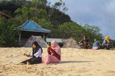 Gunungkidul, Yogyakarta, Endonezya - 24 Eylül 2020: Kapüşonlu iki kadın Pok Tunggal 'ın beyaz kumlu sahilinde oturmuş ziyaretçilerin fotoğraf çekmesini bekliyor.