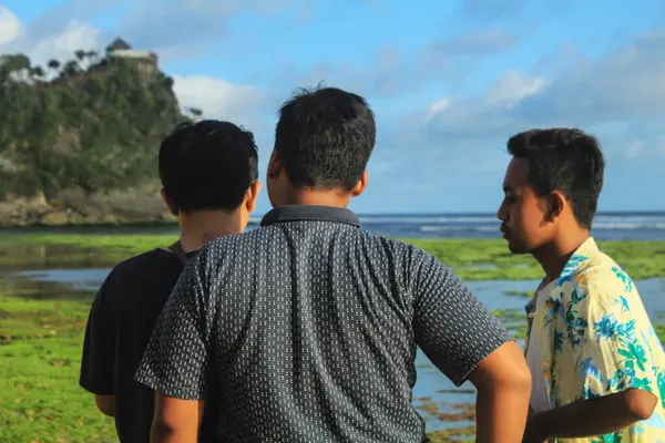Gunungkidul, Yogyakarta, Endonezya - 24 Eylül 2020: Bazı ziyaretçiler deniz bitkileriyle kaplanmış çakıllı beyaz kum ve mercan kayalarıyla dolu kıyıdan Pok Tunggal Sahili manzarasının keyfini çıkarıyorlar.