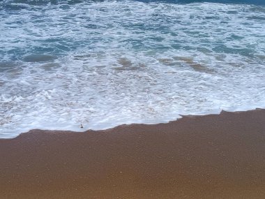 Köpüklü okyanus dalgaları yavaşça kumlu bir sahile vuruyor, rahatlatıcı ve huzurlu bir sahil manzarası yaratıyor..