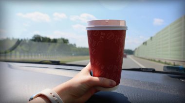 Holding a Takeaway Coffee Cup in a Car on a Road Trip. A hand holding a red takeaway coffee cup on a car dashboard while driving on a highway. The sunny weather and clear blue sky create a perfect road trip atmosphere. clipart