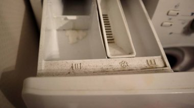Çamaşır makinesindeki kirli deterjan çekmecesinin yakın plan görüntüsü. Yıkanma öncesi, ana yıkama ve kumaş yumuşatıcısı için kompartımanlar görülebiliyor. Kirlilik ve deterjan kalıntıları birikiyor..