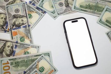 Temiz beyaz bir yüzey üzerine dağılmış yüz dolarlık banknotlarla çevrili akıllı telefon, iş, banka, pazarlama, reklam için.