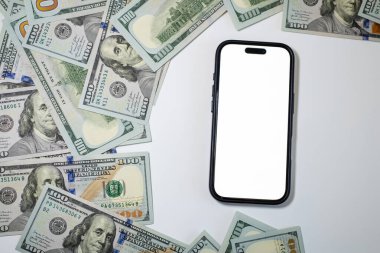 Beyaz ekranlı akıllı telefon, temiz beyaz bir yüzeye dağılmış yüz dolarlık banknotlarla çevrili. Bu da zenginlik ve finans temalarını ortaya koyuyor..