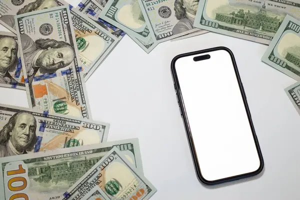Temiz beyaz bir yüzey üzerine dağılmış yüz dolarlık banknotlarla çevrili akıllı telefon, iş, banka, pazarlama, reklam için.