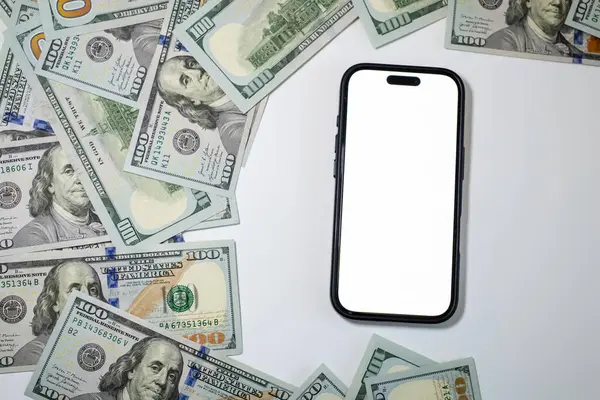 Beyaz ekranlı akıllı telefon, temiz beyaz bir yüzeye dağılmış yüz dolarlık banknotlarla çevrili. Bu da zenginlik ve finans temalarını ortaya koyuyor..
