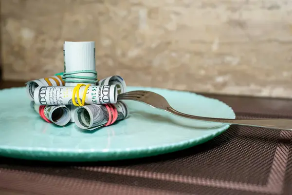 Plaka düzgünce katlanmış dolar banknotlarıyla plastik bir bantla bağlanmış, bu da parlak bir görüntü oluşturuyor. Yanında bir çatal ve bıçak duruyor. Yemek, gıda ve fiyat artışları öneriliyor..