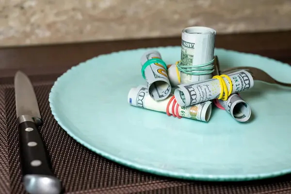 Plaka düzgünce katlanmış dolar banknotlarıyla plastik bir bantla bağlanmış, bu da parlak bir görüntü oluşturuyor. Yanında bir çatal ve bıçak var..