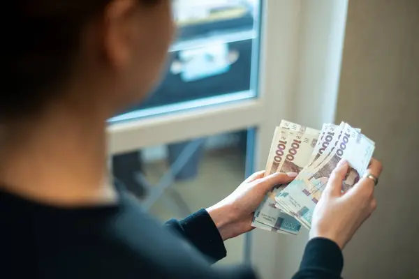 Kadın, pencerenin yanındaki parlak bir odada dururken çeşitli banknotlar tutuyor. Nakit para, dikkatle düzenlediği gibi ellerinde görünüyor. Bu da finansal yönetim anını gösteriyor. Ukrayna Hryvnia 'sı
