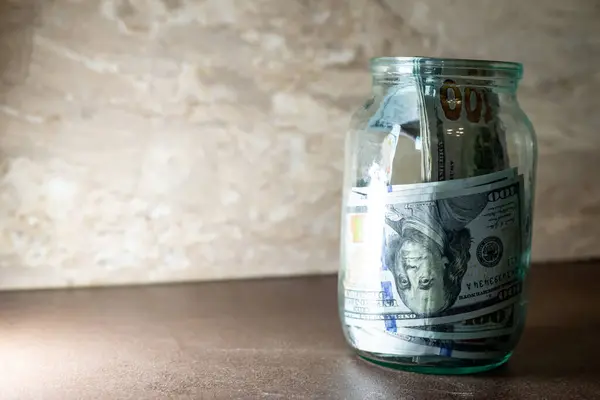 Birkaç yüz dolarlık banknotlarla dolu cam kavanoz pürüzsüz bir ahşap yüzey üzerinde duruyor. Yumuşak ışık, minimalist alanda huzurlu bir atmosfer yaratarak parayı aydınlatır. Birikmeler.