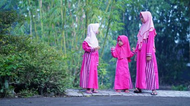 Arka planda bambu ağacı olan bir köyde pembe kıyafetler giyen ve başörtüsü takan üç kız. Seçilene odaklan