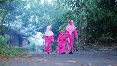 Arka planda bambu ağacı olan bir köyde pembe kıyafetler giyen ve başörtüsü takan üç kız. Seçilene odaklan