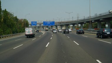 Gündüz vakti Jakarta şehrinde otoyol. Bir sürü araba ve tabela Halim PK Havaalanı, Cawang, Jagorawi, Grogol, Priok 'u gösteriyor.