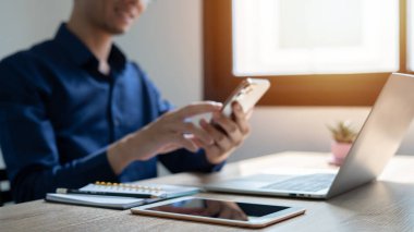 Bir adam dizüstü bilgisayarı ve cep telefonu olan bir masada oturuyor. Gülümsüyor ve eğleniyor.