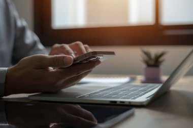 Bir kişi dizüstü bilgisayar ve saksı bitkisiyle masada otururken cep telefonu kullanıyor. Verimlilik ve odaklanma kavramı, kişinin kendi cihazları üzerinde işi veya iletişimi olduğu için