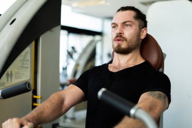 Spor salonunda bir adam bir makinenin üzerinde oturuyor. Siyah bir gömlek giyiyor ve sakalı var.