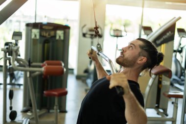 Bir adam spor salonunda ağırlık kaldırıyor. Siyah bir gömlek giyiyor ve sakalı var.