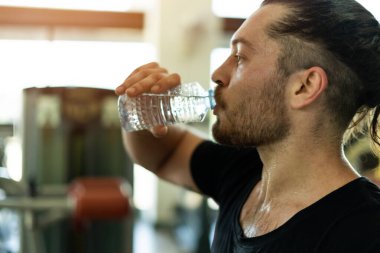 Egzersiz yaparken bir adam şişeden su içiyor. Şişe plastikten yapılmış ve suyla dolu. Adam siyah bir gömlek giyiyor ve sakalı var.