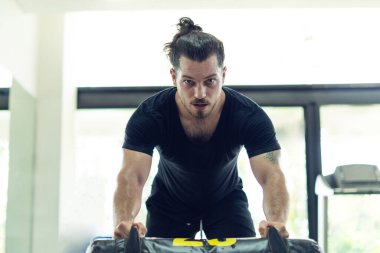 Bir adam spor salonunda ağırlık kaldırıyor. Siyah bir gömlek ve siyah bir şort giyiyor.