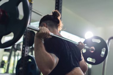 Bir adam spor salonunda ağırlık kaldırıyor. Sahne odaklanmış ve odaklanmış. Adam bir halter kaldırır ve çömelir. Bu da gücünü ve formülünü geliştirmek için çok çalıştığını gösterir..