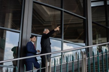 Takım elbiseli iki adam korkulukların üzerinde duruyor, biri bir şeyi işaret ediyor. Sahne profesyonel ve ciddidir.