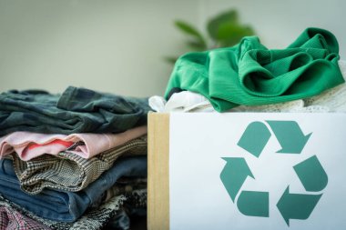 Yeşil geri dönüşüm sembolü olan bir kutunun üstüne bir kutu dolusu kıyafet yığılır. Kıyafetlerin hepsi farklı renkler ve stiller, ve kutu onlarla dolup taşıyor. Giysileri geri dönüşüm ve yeniden kullanma kavramı