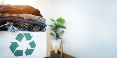 Üzerinde yeşil geri dönüşüm işareti olan bir kutu giysilerle doludur. Arka planda saksı bitkisi var.