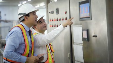 Güvenlik teçhizatlı iki adam kontrol panelini işaret ediyor. Sahne ciddi ve odaklı.