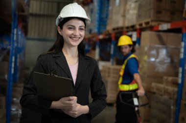 Sert şapka takan ve elinde dosya tutan bir kadın. Arkasında çelik yelekli bir adam var.