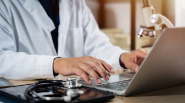 Bir doktor, tıbbi bir ortamda dizüstü bilgisayarda yazı yazıyor. Laptop açık ve doktor bunu hasta bilgilerine ulaşmak ya da diğer tıp uzmanlarıyla iletişim kurmak için kullanıyor.