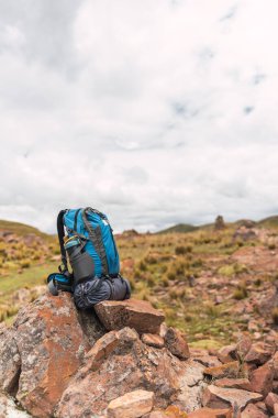 Dağdaki kayalara yerleştirilmiş mavi yürüyüş çantası yeşil bitki örtüsü ve güneş ışığı ve fırtına bulutlarıyla çevrili.
