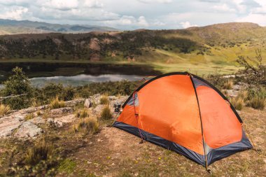 Bir dağın tepesinde turuncu bir çadır, arka planda bir göl ve mavi gökyüzü ve And Dağları 'nın ortasında ormanın ortasında bulutlar.