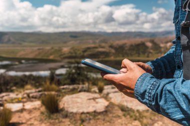Yeşil bitki örtüsü ve doğal güneş ışığı ile çevrili bir cep telefonu tutan mavi gömlekli eller ve And Dağları 'nın arka planında bir orman olan fırtına bulutları.
