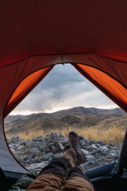 Adamın ayakları, dağlardaki turuncu çadırın içinde gün batımında kayalar ve sarı bitki örtüsüyle çevrili kışın And Dağları sıradağlarında