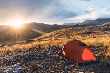 Dağlarda turuncu renkli kamp. Gün batımında. Kayalar ve sarı bitki örtüsüyle çevrili.
