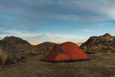Dağlarda turuncu renkli kamp. Gün batımında. Kayalar ve sarı bitki örtüsüyle çevrili.