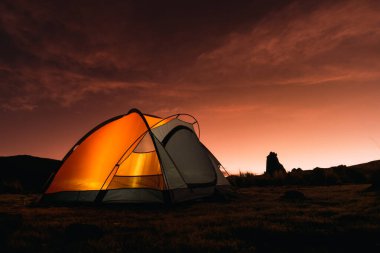 Dağlarda turuncu renkli kamp. Gün batımında. Kayalar ve sarı bitki örtüsüyle çevrili.