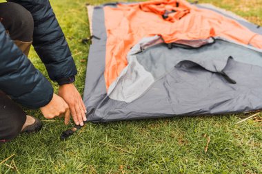 Adamın elleri, And Dağları 'nın doğal ışıklarıyla aydınlanan yeşil çimlerin üzerine turuncu bir çadır kuruyor.