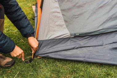 Adamın elleri, And Dağları 'nın doğal ışıklarıyla aydınlanan yeşil çimlerin üzerine turuncu bir çadır kuruyor.