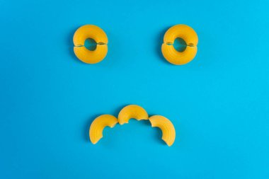 Mavi bir yüzey üzerinde ahşap bir kaşıkta erişteler ve mutlu ve hüzünlü ifadeleri olan bir doğrama tahtası.