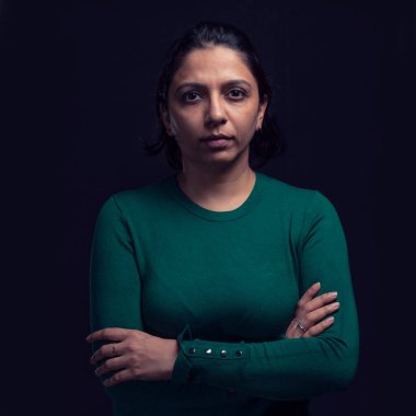 Güzel, ciddi, gizemli, yeşil giysili bir kadın karanlıkta kollarını kavuşturmuş dramatik bir şekilde duruyor. Karanlık ve sade bir portre. - Sanat. Siyah arkaplanda ön yüz görünümünü kapat. 