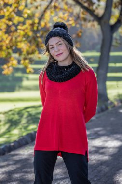 Beautiful young blonde woman, donning a black woolen hat and a vibrant red sweater, stands enchantingly beneath the autumn trees clipart