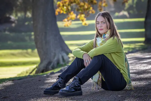 stock image A seated young blonde woman dressed in cozy attire sits beneath autumn trees, her arms casually resting on her knees