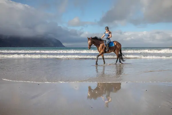 stock image Teenage girl cantering brown horse along shoreline at beach in sunshine with beautiful sea in background