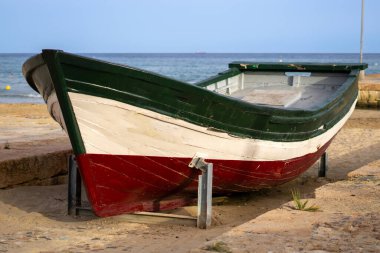 İspanya, Salou 'da kumlu sahilde eski ahşap bir tekne. Tatilciler ve arka planda cankurtaranlar var. Bulutlu mavi gökyüzü.